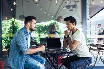 Side view of Caucasian male colleagues sitting at cafeteria table and discussing web project and graphic design during collaborative meeting for brainstorming on ideas, skilled IT professionals