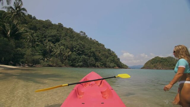 Young woman in sunglasses pulls pink plastic canoe with paddle to azure sea water from beach with palms.