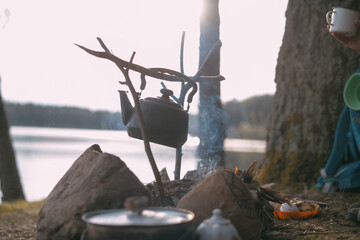 A camping kettle hangs over a campfire on the shore of a lake on a camping trip. Field kitchen by...