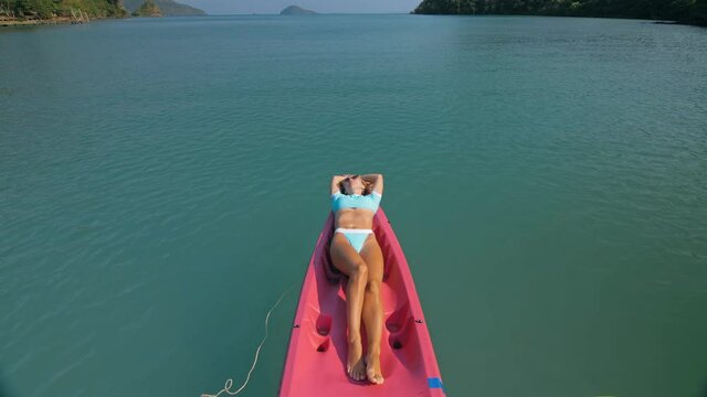 Pretty slim woman in elegant swimsuit lies in pink plastic kayak drifting on azure ocean at exotic resort upper view.