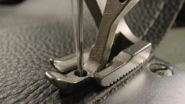 Macro shot of sewing machine needle with thread and presser foot tailoring leather piece during sewing process in handmade workshop. Slow motion of production operation. Sewing equipment close up.