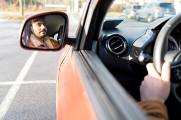 Hispanic or eastern ethnicity man holding driving wheel riding car on parking slot in morning. Travel, exam, lesson, learning, taxi driver. View from outside, opened window, face in rearview mirror