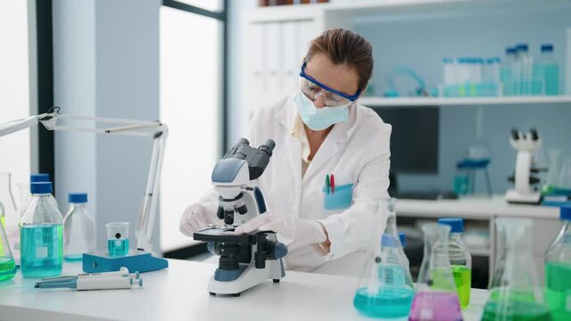 Middle age woman wearing scientist uniform and medical mask using microscope at laboratory