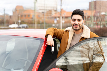Fototapeta na wymiar Young latin or arab eastern indian pakistani smiling man holding ignition key standing near red new clean bought car with opened door on parking slot of urban city. Travel, rental, car sharing