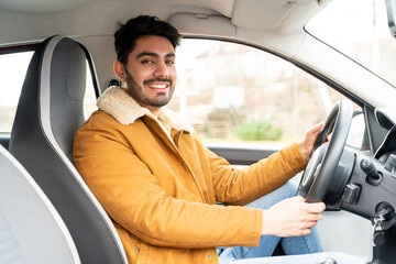Portrait of smiling spanish or arab eastern ethnicity man casually dressed with hands holding driving wheel in car on sunny autumn, winter or spring day. Travel, exam, lesson, learning, taxi driver