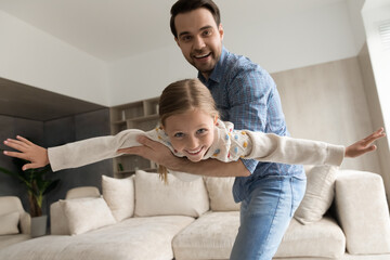 Portrait of happy caring young dad holding in hands flying joyful adorable laughing child daughter, enjoying domestic entertaining weekend activity, playing games at home, good relations concept.