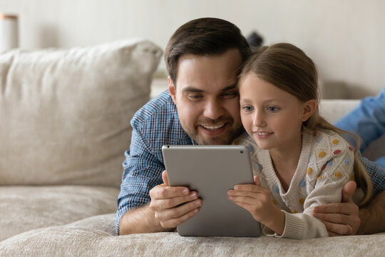 Smiling loving millennial father embracing cute small child daughter, enjoying using interesting software application on digital computer tablet, lying together on sofa, copy space for advertisement.