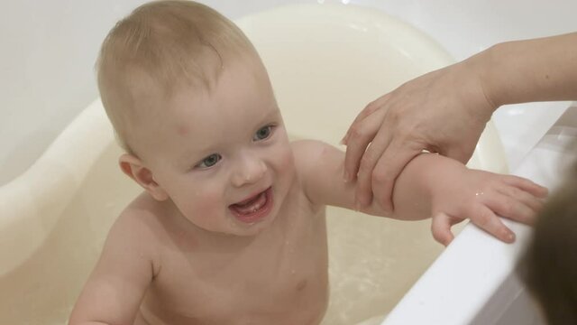 Mother with cute little child have fun while bathing kid in baby bath, 10 month old baby boy smiling playing in clean water in the bath closeup. High quality 4k footage