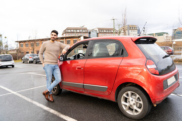 Young latin or arab eastern indian pakistani carefree man in stylish clothes standing near red small car on parking slot of an urban city background on rainy midseason day. Travel, rental, car sharing