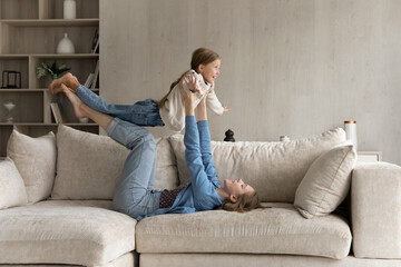Happy young mother lying on cozy sofa, lifting in air joyful laughing small child daughter, playing airplane game, imagining travelling or practicing balance exercise, having fun together at home.