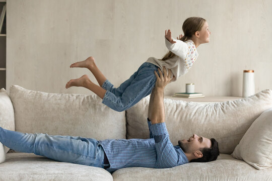 Joyful young 30s father lying on couch, lifting in arms in air laughing adorable preteen small kid daughter, practicing balance, playing airplane game, imagining traveling, having fun together at home
