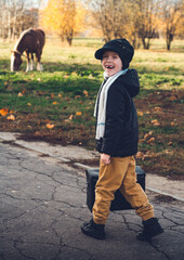 Fototapeta premium A cute little traveler without front teeth smiles, he holds a large leather bag in his hand, steps towards his horse, which is grazing nearby.