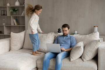 Concentrated young father working remotely on computer, sitting on sofa while small energetic happy child daughter jumping and needs attention, single parenting problems, quarantine lifestyle concept.