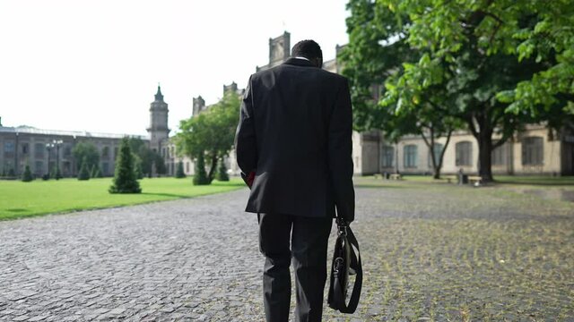 Tracking shot African American professor in suit walking on university yard in the morning. Back view confident intelligent young man strolling to college building outdoors. Education and profession