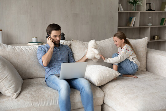 Focused Young Father Multitasking At Home, Holding Cellphone Conversation Working On Computer And Playing Toys With Cute Small Kid Daughter, Sitting Together On Comfortable Sofa, Quarantine Lifestyle.