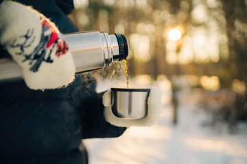 Woman pours hot tea from a thermos into a mug, walking in snowy frozen winter forest. Adventure, travel, tourism and camping concept