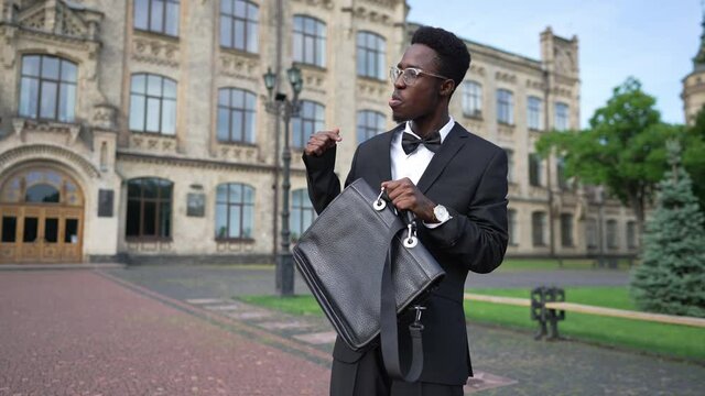 Medium shot portrait of self assured elegant African American man in suit with attache case standing at college campus outdoors talking gesturing. Happy new teacher at university in the morning