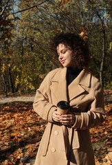 Curly hair woman in beige coat with cup of coffee outdoor. Beautiful young woman drinks coffee in the autumn park with falling foliage.