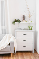 Beautiful plates, glasses with fir branches and pine cones on chest of drawers near white wall