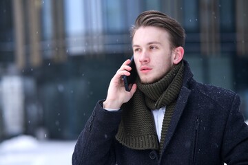 Portrait of serious handsome man, businessman, business person is talking on his cell mobile phone, calling on smartphone outdoors at winter cold snowy day in snow in coat scarf, having conversation