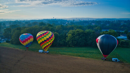 Fototapeta premium Multiple Hot Air Balloons Landing in Farmlands as Amish Look on, and More Still in Flight