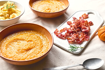 Pumpkin cream soup with roasted bacon and croutons in clay plate on the table with linen tablecloth. Autumn vegetable creamy soup puree. Healthy seasonal eating.
