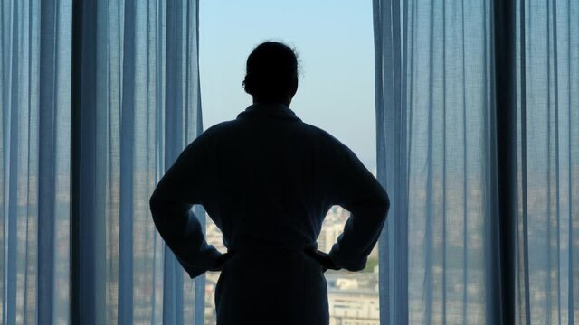 Sleep-deprived traveler goes to window and looks out over city, standing in bath robe with his hands on hips. After standing motionless, he turns around and walks off into dark of the room
