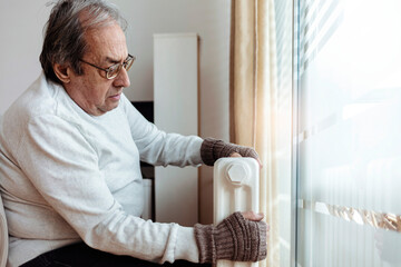 Cropped shot of a senior men in woolen gloves and a sweater warms his hands near the heater. An older man is sitting near a radiator at home, wearing gloves and warming his hands.