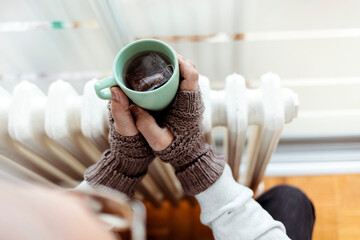 A cold grandfather wearing gloves, warming himself by the radiator and drinking hot tea at home....