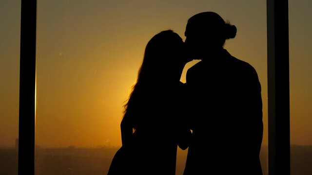 Woman and man hold a small toy house, woman reaches out to man and kisses him. Happy couple stand against window, silhouetted shot, sunset sun shine through little house
