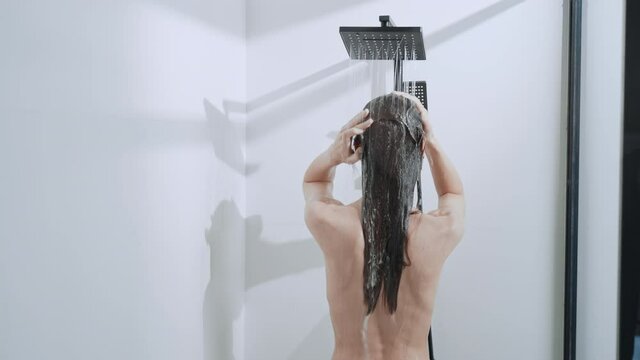 rear view caucasian woman washing long hair with shampoo in shower cabin splashing water flow down the body
