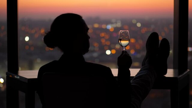 Man sits alone in a dark room by a window, stir and slightly shaking glass of white wine in hand. Lazy evening alone after a busy day at work. Dark city and lights seen blurred on background