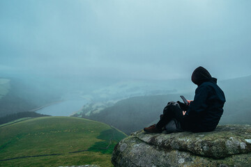 Fototapeta premium Young male in dark clothes sitting on a rock formation edge looking at phone when surrounded by beautiful landscape. Mobile addiction issue outdoors. Hooded man wearing black browsing on cellphone.