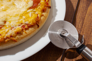 Cooked cheddar cheese pizza on a white plate and stainless steel pizza cutter dark brown wooden table. Italian style dish. Fast food industry product. Close up