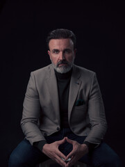 Portrait of adult businessman wearing trendy suit and sitting in modern studio on stylish chair against the black background