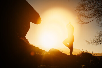 silhouette of man practicing yoga, performing the tree pose on a rock in the mountain at sunset. healthy life style