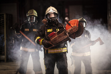 Fototapeta premium Group of professional firefighters wearing full equipment, oxygen masks, and emergency rescue tools, circular hydraulic and gas saw, axe, and sledge hammer. smoke and fire trucks in the background.