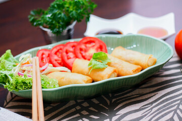 Fried spring rolls, vegetables and tomatoes placed in a green leaf shape plate on a black wooden table and dipping sauce.