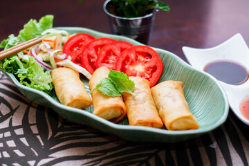 Fried spring rolls, vegetables and tomatoes placed in a green leaf shape plate on a black wooden table and dipping sauce.