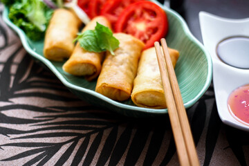 Fried spring rolls, vegetables and tomatoes placed in a green leaf shape plate on a black wooden table and dipping sauce.