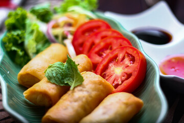 Fried spring rolls, vegetables and tomatoes placed in a green leaf shape plate on a black wooden table and dipping sauce.