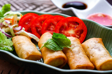 Fried spring rolls, vegetables and tomatoes placed in a green leaf shape plate on a black wooden table and dipping sauce.