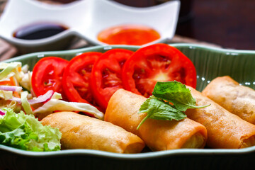 Fried spring rolls, vegetables and tomatoes placed in a green leaf shape plate on a black wooden table and dipping sauce.