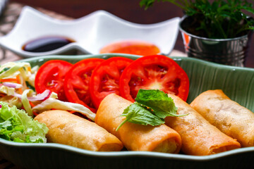 Fried spring rolls, vegetables and tomatoes placed in a green leaf shape plate on a black wooden table and dipping sauce.