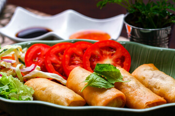 Fried spring rolls, vegetables and tomatoes placed in a green leaf shape plate on a black wooden table and dipping sauce.