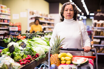 Pensive middle aged female shopping at store, walking among shelves and choosing products