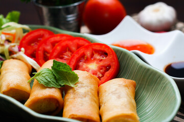 Fried spring rolls, vegetables and tomatoes placed in a green leaf shape plate on a black wooden table and dipping sauce.