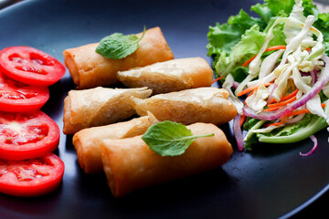 Fried spring rolls with vegetables and tomatoes placed in a black plate on a black wooden table and dipping sauce.