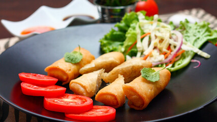 Fried spring rolls with vegetables and tomatoes placed in a black plate on a black wooden table and dipping sauce.