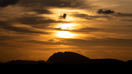 Silhouette of a hill against beautiful golden hour light and beautiful clouds formation in the horizon with the sun setting

